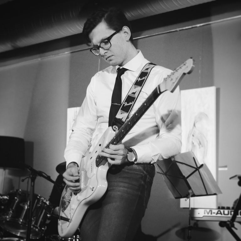 Jürgen Šuvalov playing guitar. It is a black and white image. I am wearing playing a Fender Jazzmaster. I am wearing a white shirt and black tie.  Some other musical gear such as drums and a keyboard can be seen in the background.