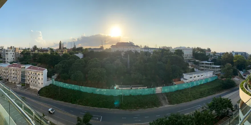 A panoramic view from the balcony of a hotel. There's a road next to the hotel, followed by a quite heavily forested area. The sun is setting behind a lonesome cloud in the background.