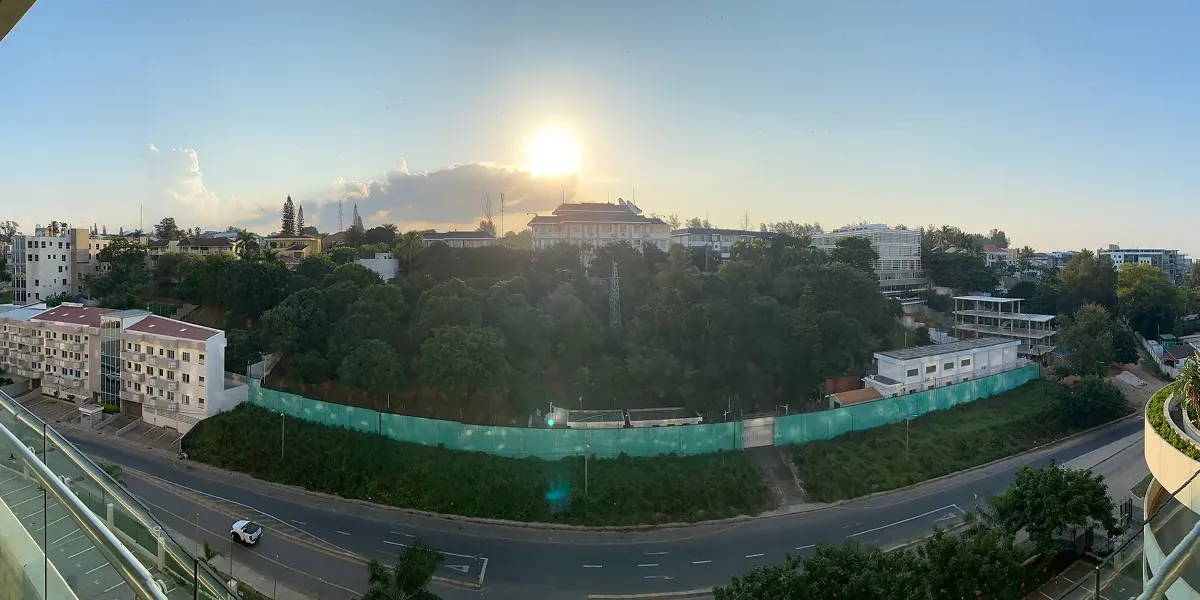 A panoramic view from the balcony of a hotel. There's a road next to the hotel, followed by a quite heavily forested area. The sun is setting behind a lonesome cloud in the background.