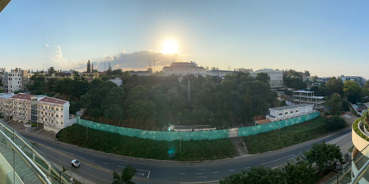 A panoramic view from the balcony of a hotel. There's a road next to the hotel, followed by a quite heavily forested area. The sun is setting behind a lonesome cloud in the background.