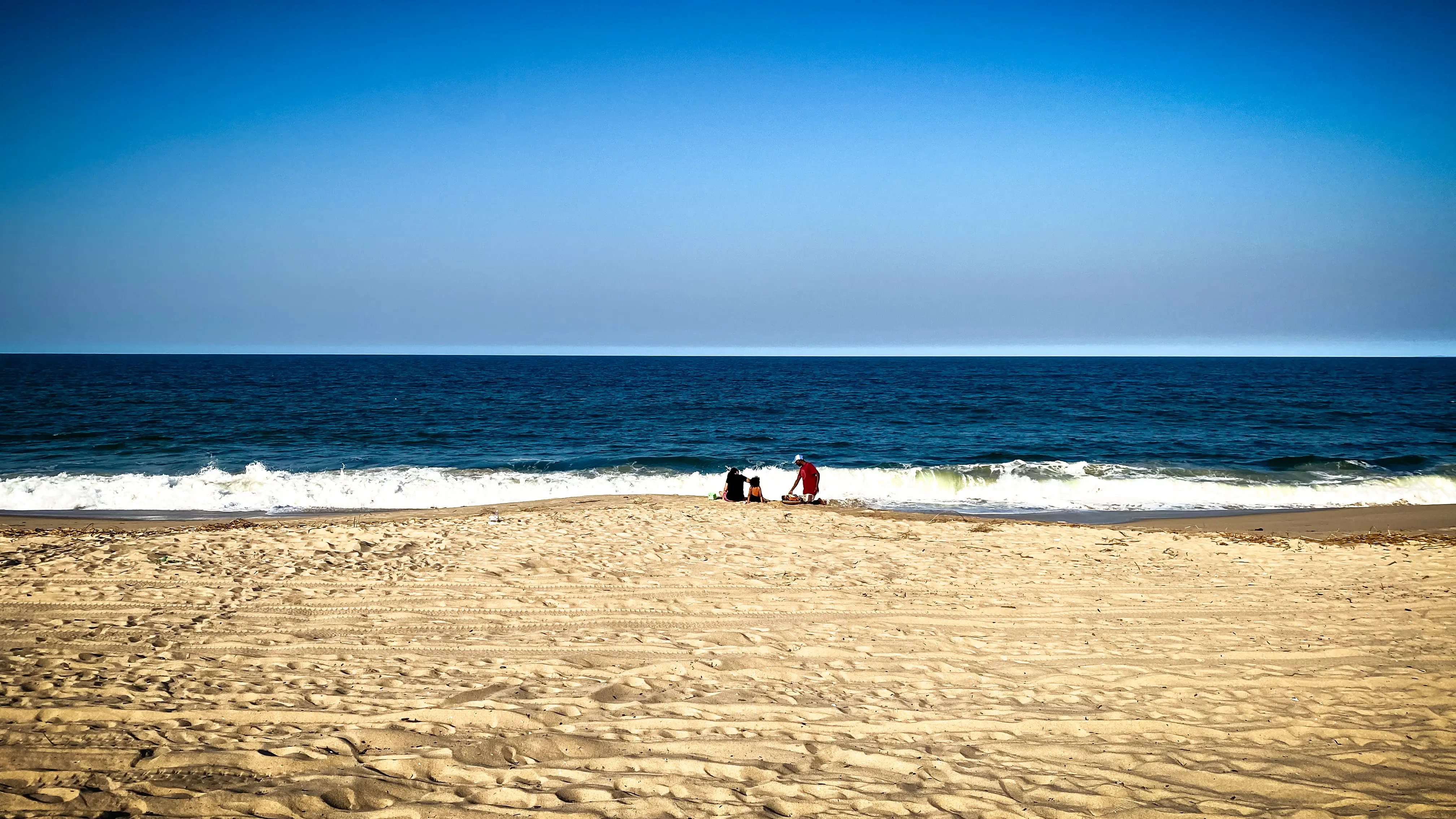 A wide, empty beach of golden sand leading down to deep blue surf under a clear sky. A few people stand near the waterline, one of them in a red shirt.