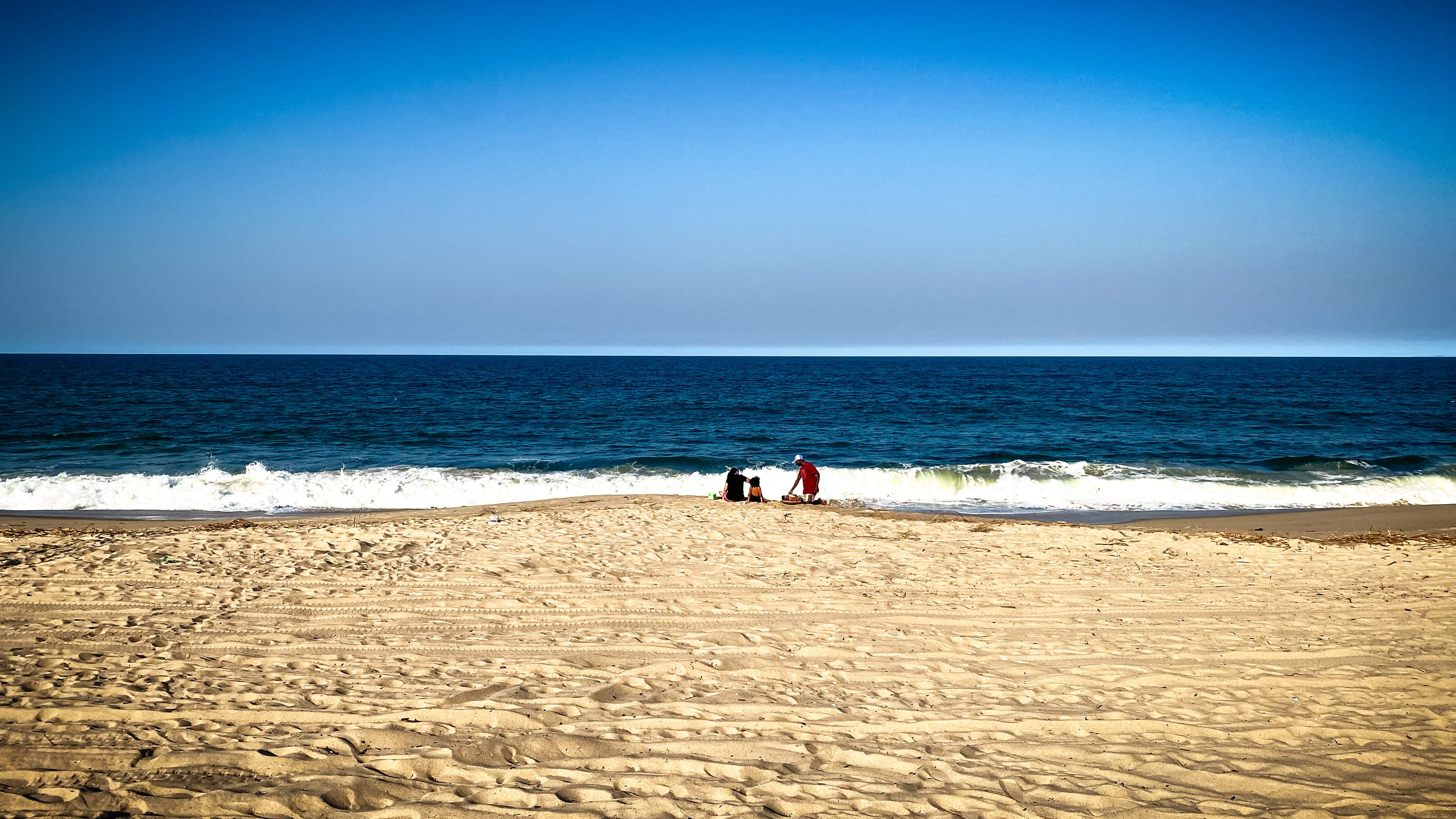 A wide, empty beach of golden sand leading down to deep blue surf under a clear sky. A few people stand near the waterline, one of them in a red shirt.
