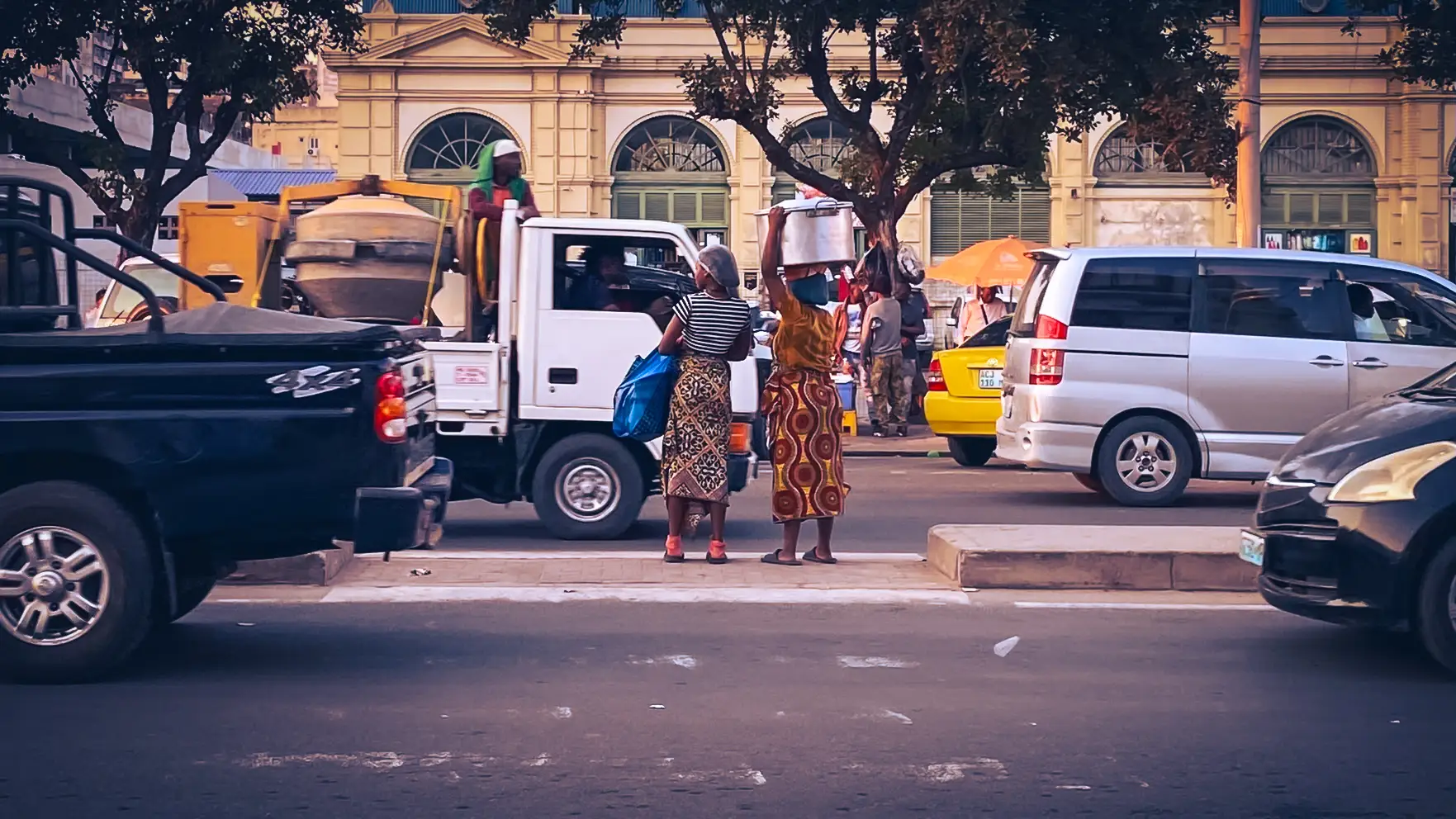 A busy Maputo street seen from across the road. Two women in patterned capulanas wait on a median, one balancing a large metal pot on her head. Cars, a pickup and a van pass by, with a grand yellow colonial-era building behind.