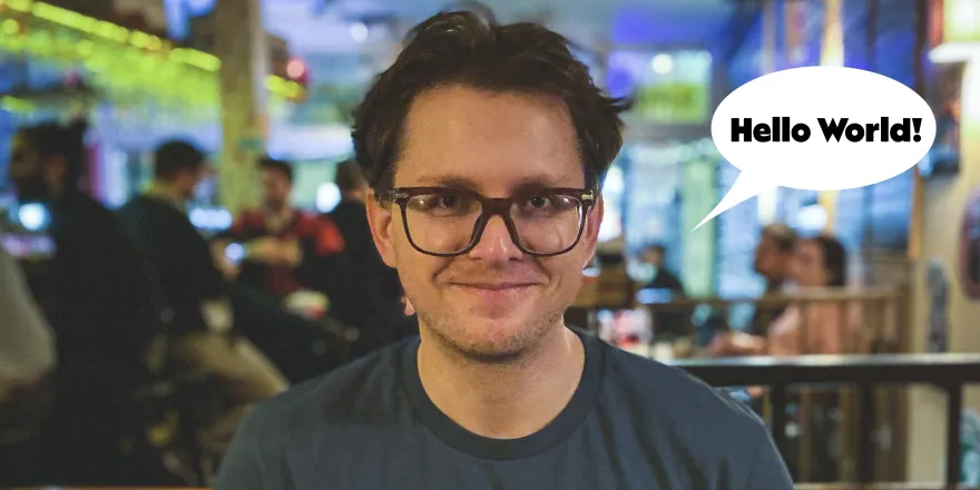 Jürgen with messy hear, grinning wide. There's a speech bubble next to his face that says "Hello World!". The background is blurred but we can see it's a bar.