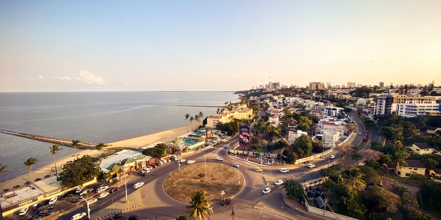 The view from my hotel room – Maputo's coastline at golden hour. A large roundabout and a palm-lined beach sit between the road and the Indian Ocean, with the city's high-rises receding into the haze along the shore.