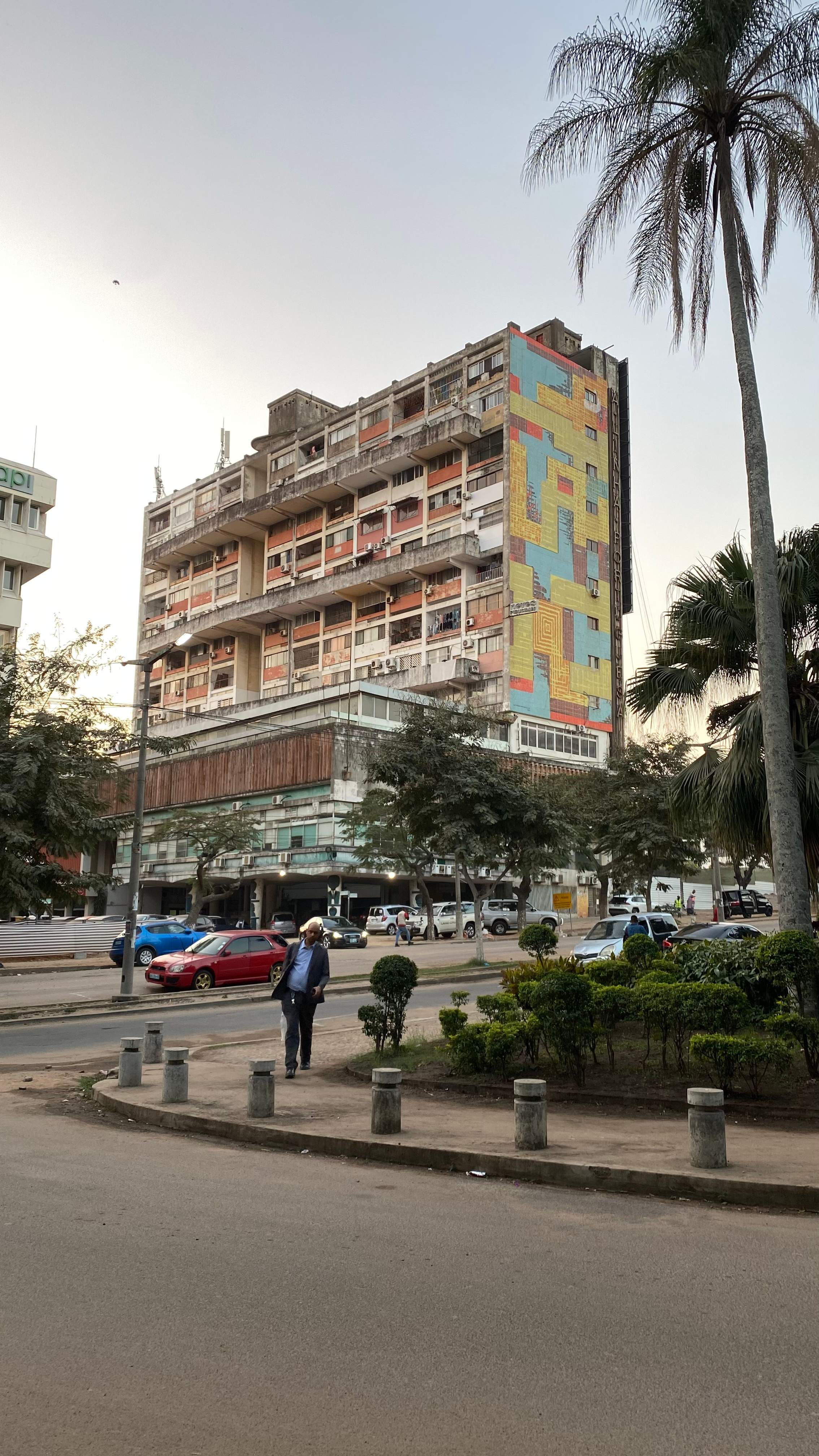 A weathered high-rise apartment block with rows of balconies, one entire side covered by a large geometric mural in blue, yellow and red. A tall palm tree stands in the foreground beside a street with parked cars.