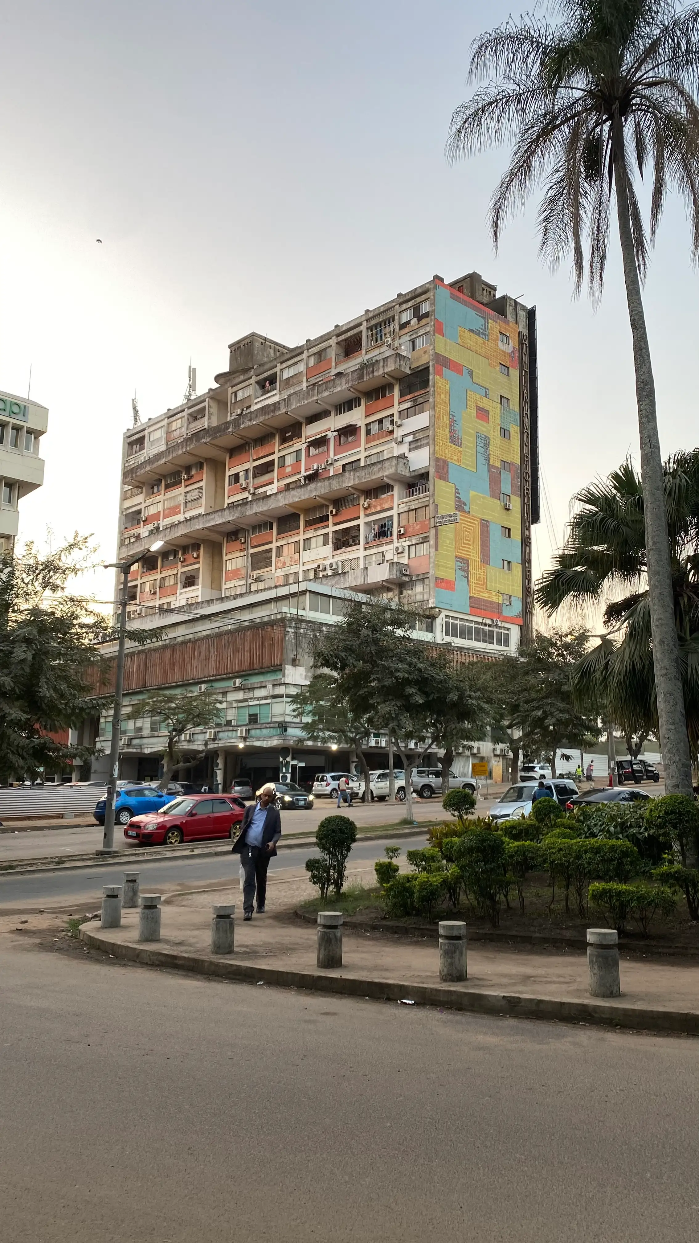 A weathered high-rise apartment block with rows of balconies, one entire side covered by a large geometric mural in blue, yellow and red. A tall palm tree stands in the foreground beside a street with parked cars.