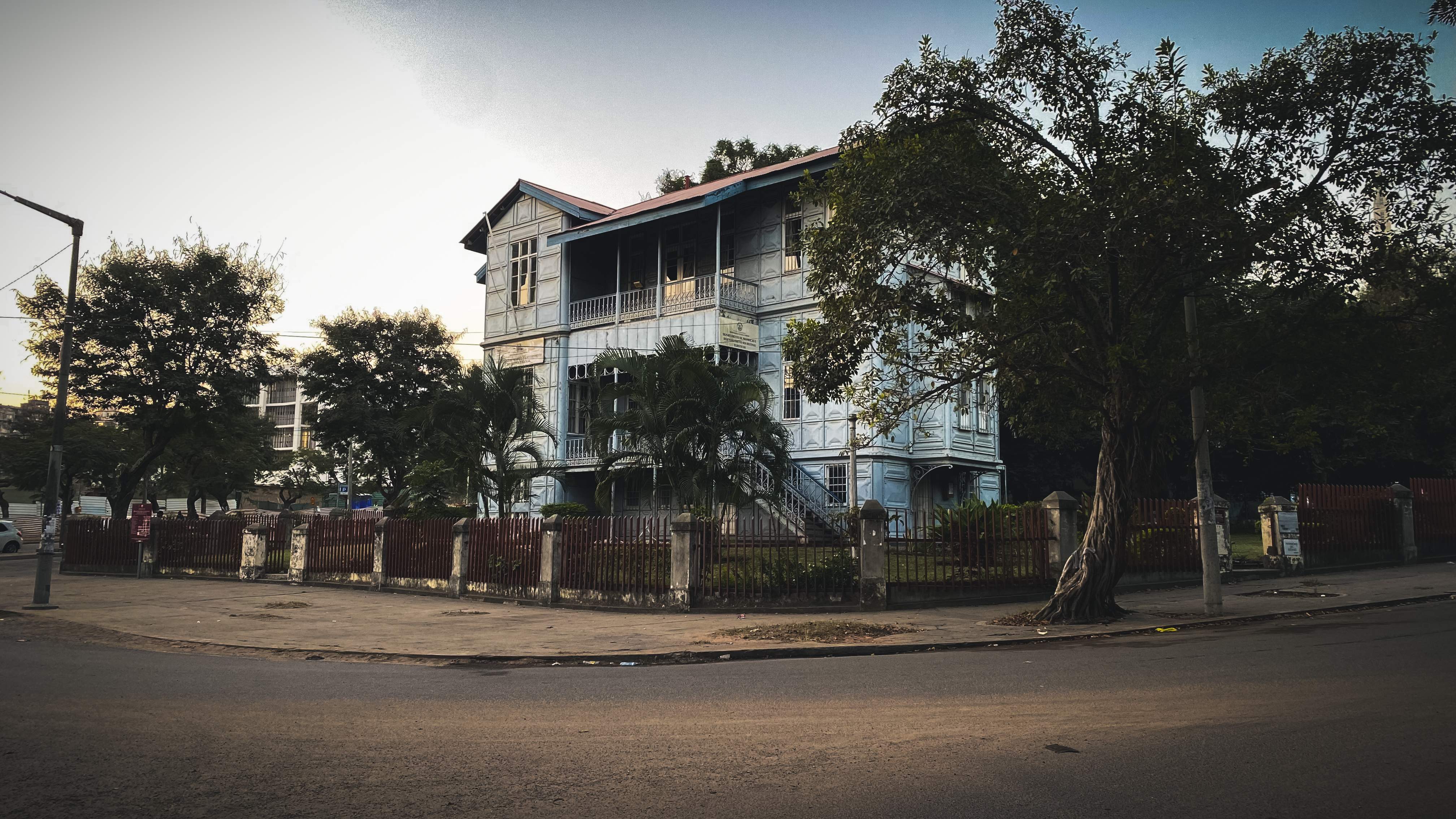 A pale blue building clad entirely in moulded metal panels, with wraparound verandas and iron railings, set behind a dark red fence and framed by trees in the evening light.