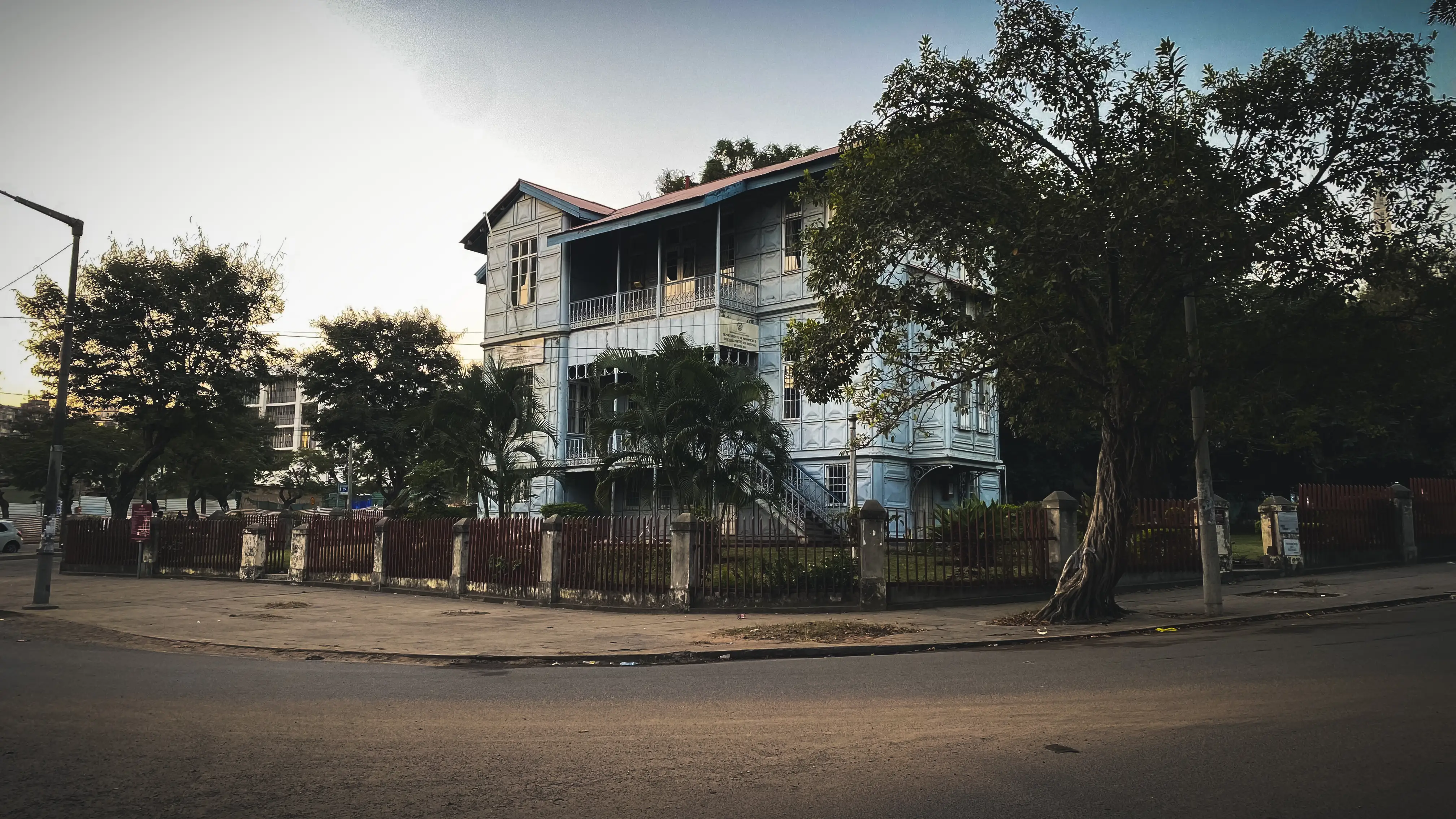 A pale blue building clad entirely in moulded metal panels, with wraparound verandas and iron railings, set behind a dark red fence and framed by trees in the evening light.