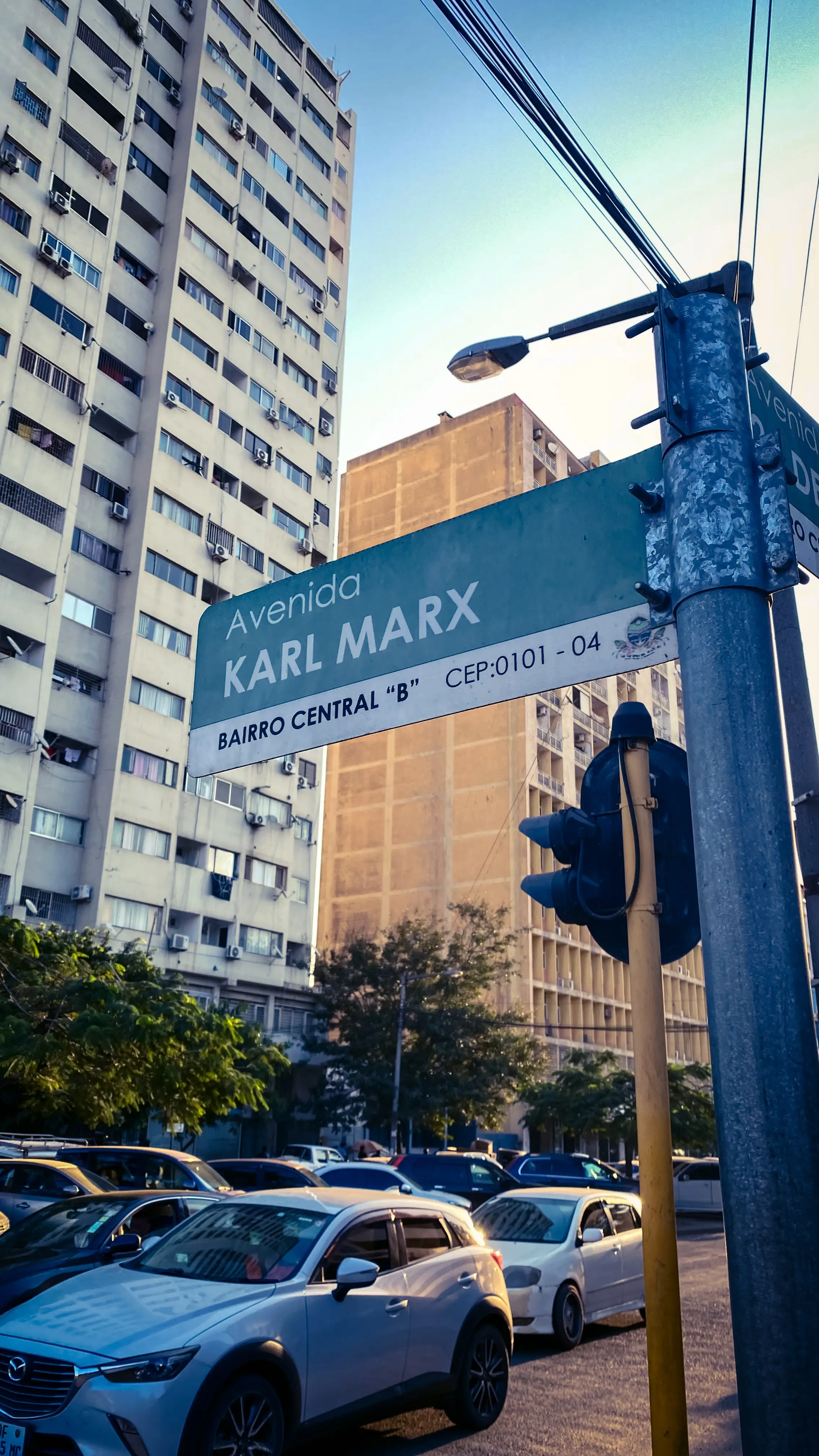 A green-and-white street sign reading 'Avenida Karl Marx, Bairro Central B', mounted on a pole with tall apartment blocks and parked cars behind it.