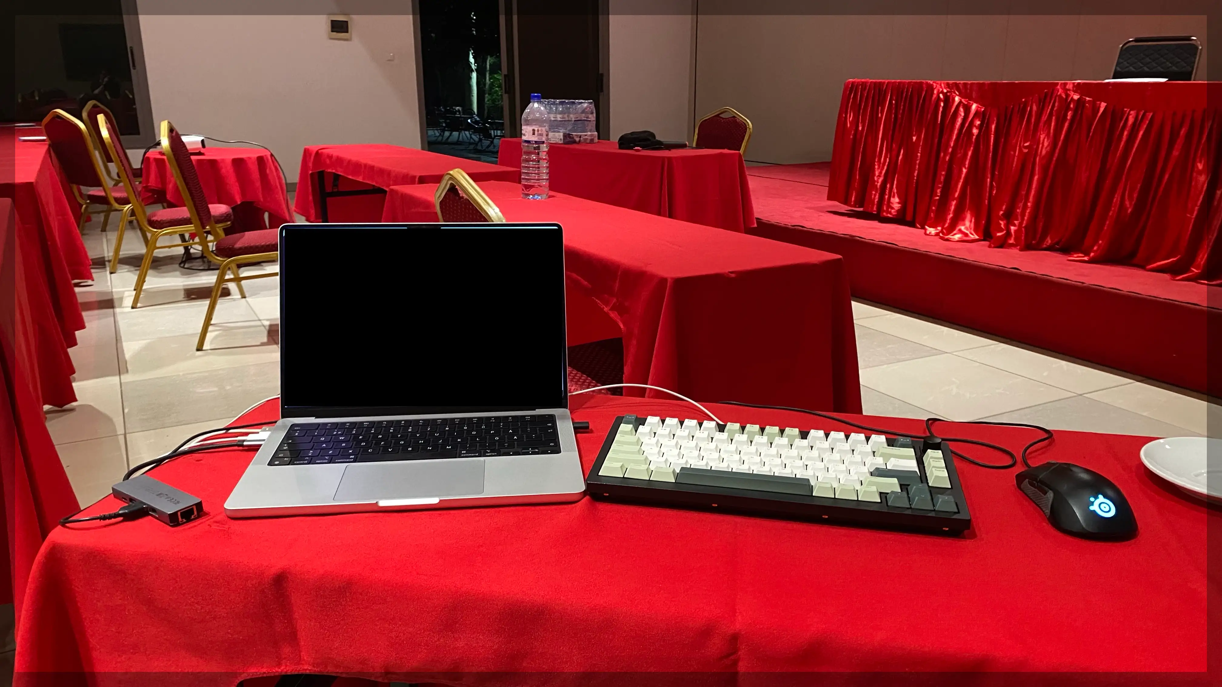 In a conference room, on table covered in red cloth, there is a Macbook laptop. Next to it a mechanical keyboard and gaming mouse. It is dark outside, seen through the door in the backkground.