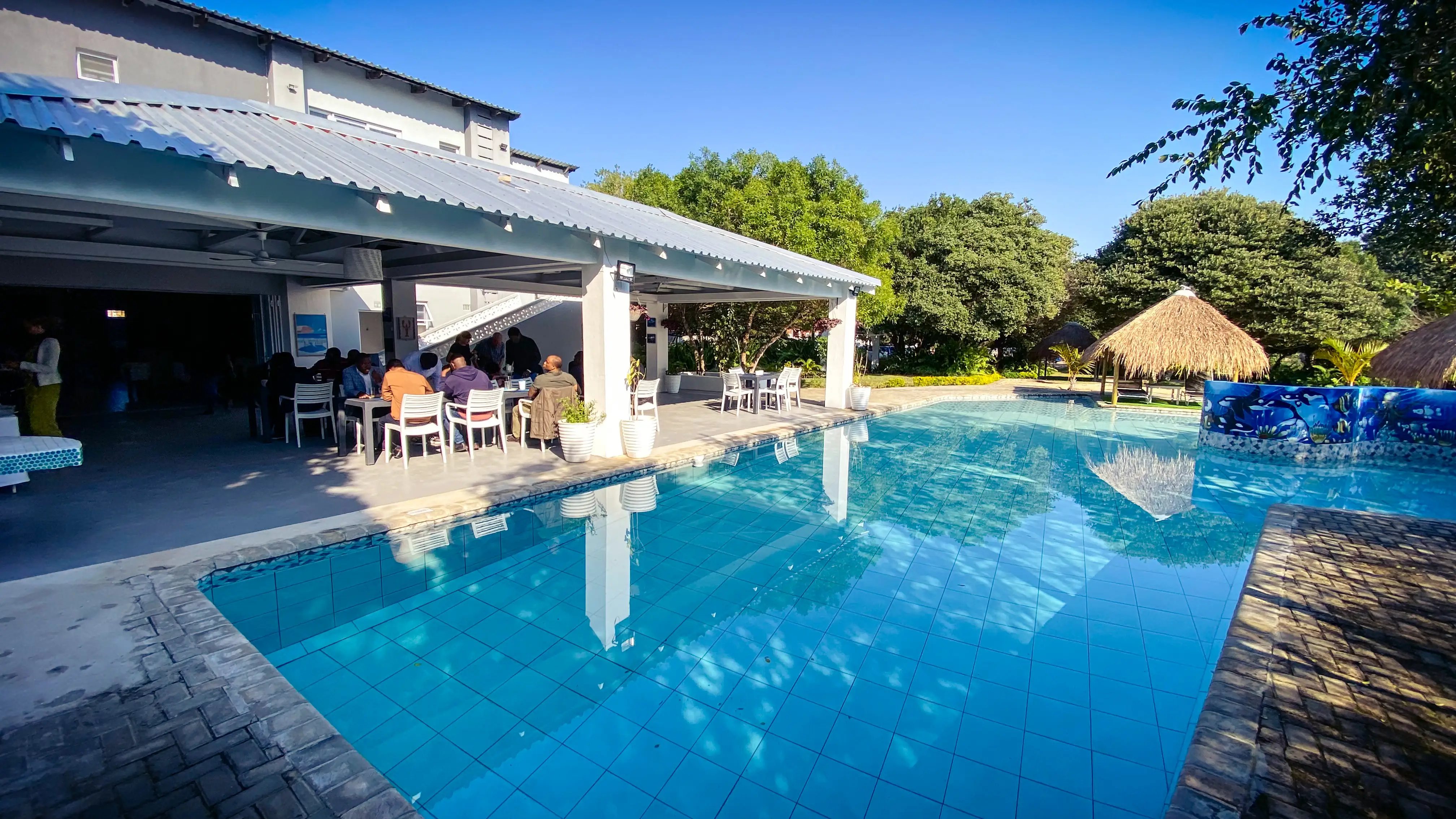 A resort swimming pool in the foreground. To the left, an open-sided building with a metal roof where people sit at white tables. A thatched umbrella, tropical trees and a clear blue sky in the background.