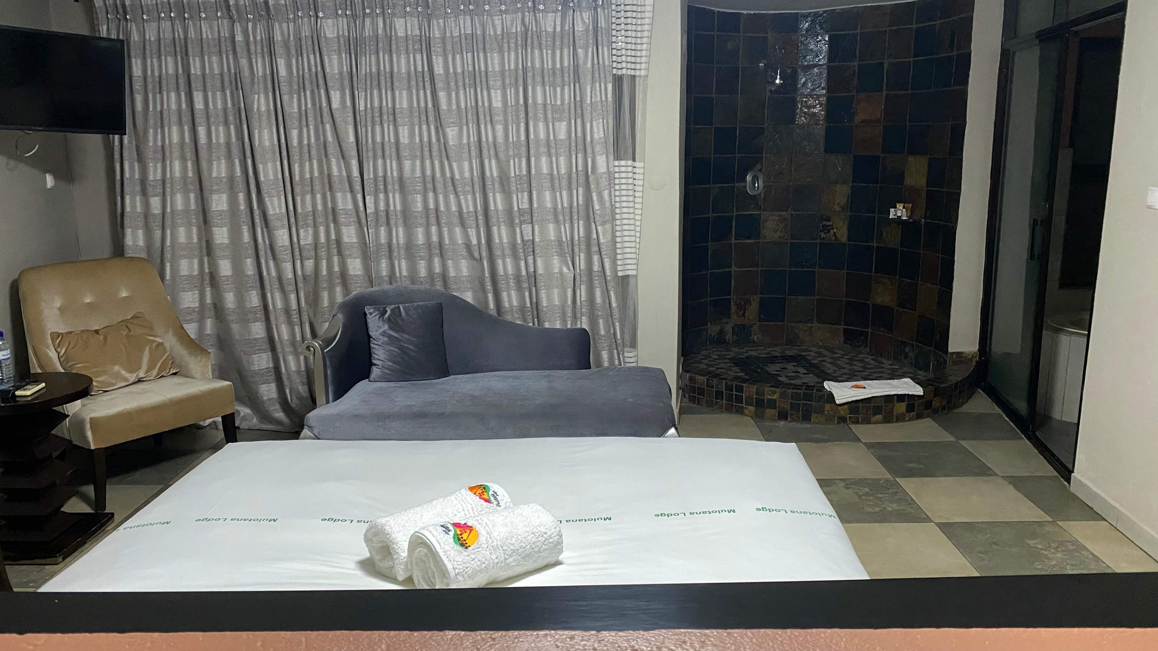 In the foreground ins a hotel bed. In the background, a chair, a sofa and in the corner an open shower with brown tiles on the wall.