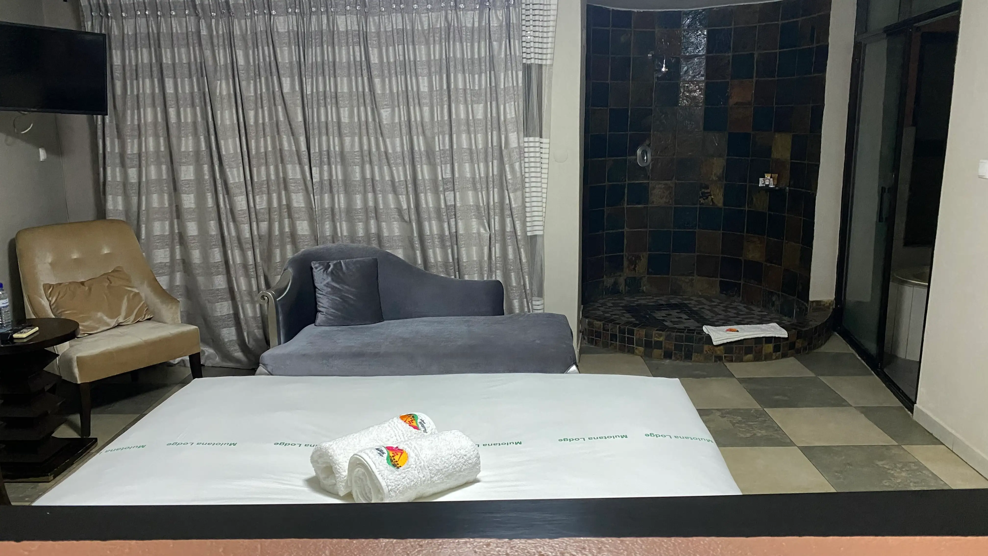 In the foreground ins a hotel bed. In the background, a chair, a sofa and in the corner an open shower with brown tiles on the wall.