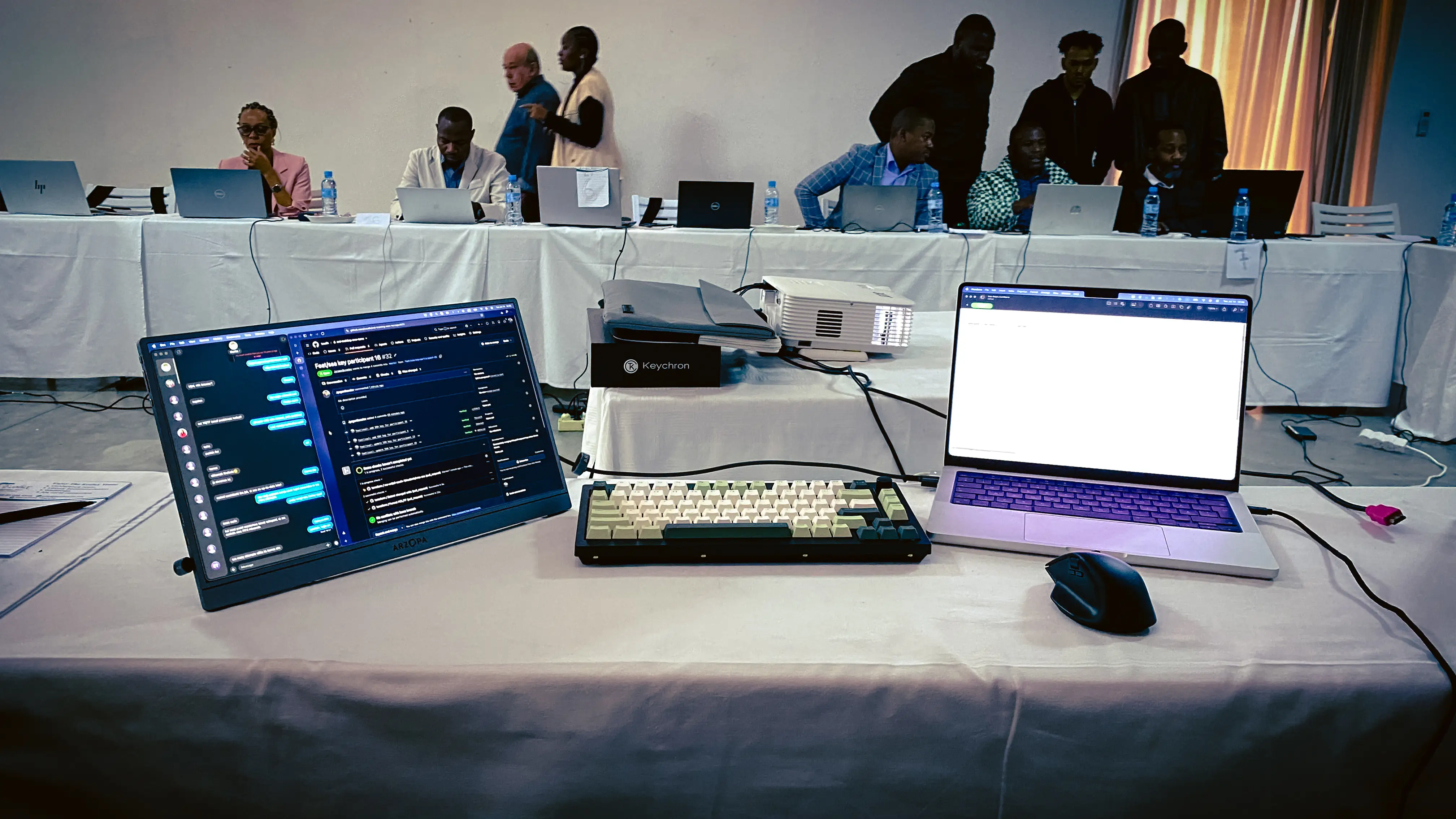 A workshop desk with a portable monitor showing a chat panel and an issue tracker on the left, a compact mechanical keyboard in the center, a MacBook with a browser open on the right, and a mouse. Behind it, participants work on laptops at cloth-covered tables.