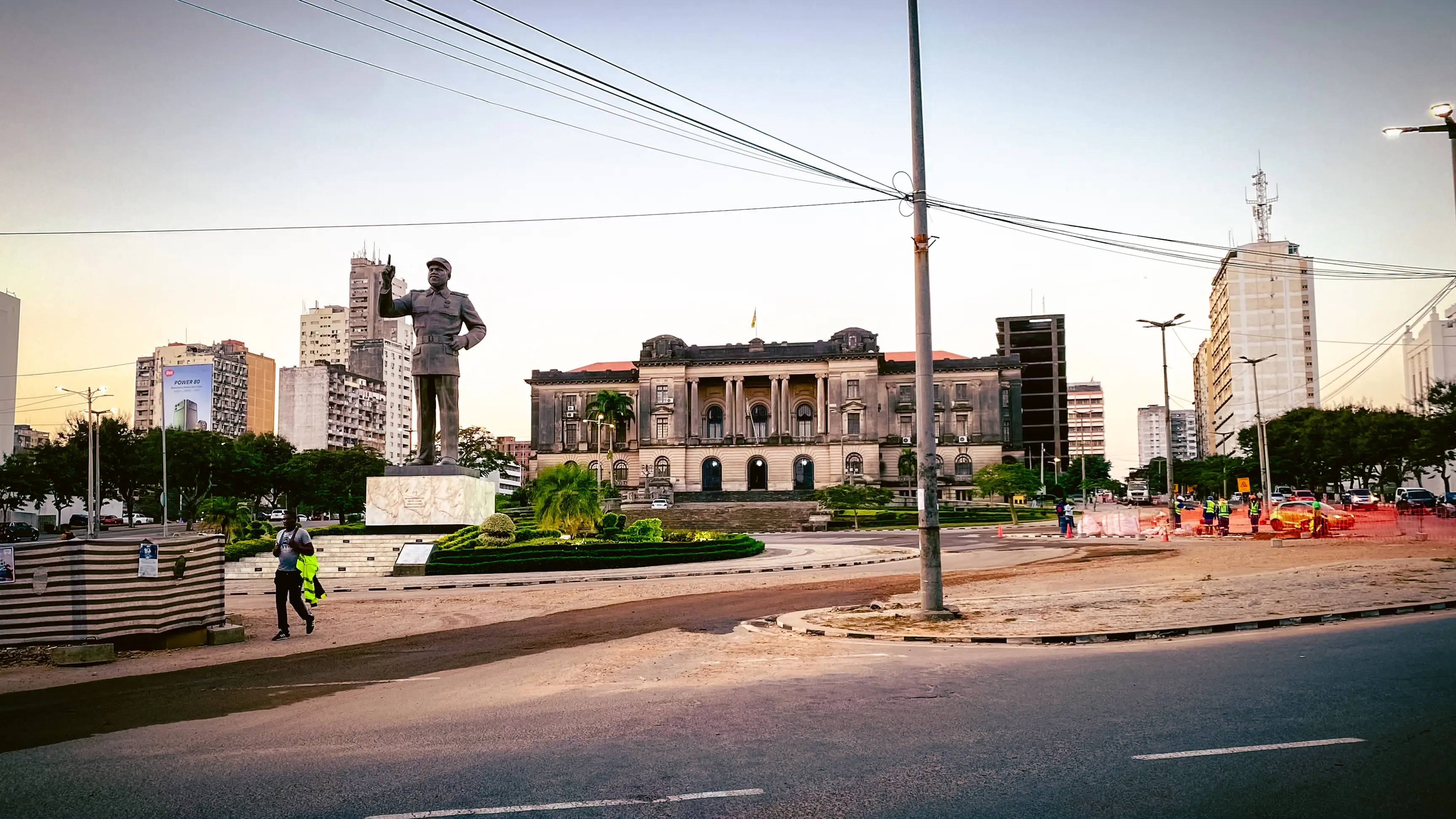 A bronze statue of a man in military uniform and cap, one finger raised, standing on a marble plinth in the middle of a roundabout. Behind it is a grand neoclassical building, with the city's high-rises around the square.